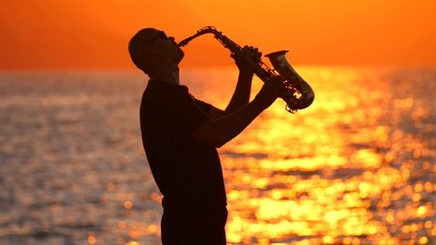 Black silhouette of young bald male saxophonist musician playing golden alt saxophone on musical instrument on seashore, amid waves, beautiful orange golden sunset, dawn. Slow motion video. Glare sun.