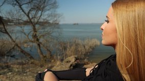 Video 4k clip, a young blonde woman with long hair sits on a red anchor car, looks at the lake on a sunny day in spring