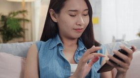 Happy Young Asian woman sits on sofa using smartphone browsing the internet or social media, watches video relaxing, and spends time at home during  quarantine. lifestyle and social distancing concept