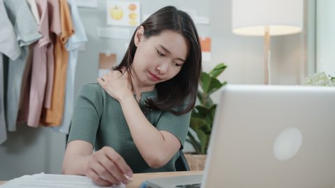 Tired Beautiful Asian Businesswoman sits at a work desk feeling fatigued, exhausted having neck pain and shoulder touching, self-massaging, and stretching with a painful face. Office Syndrome Concept.