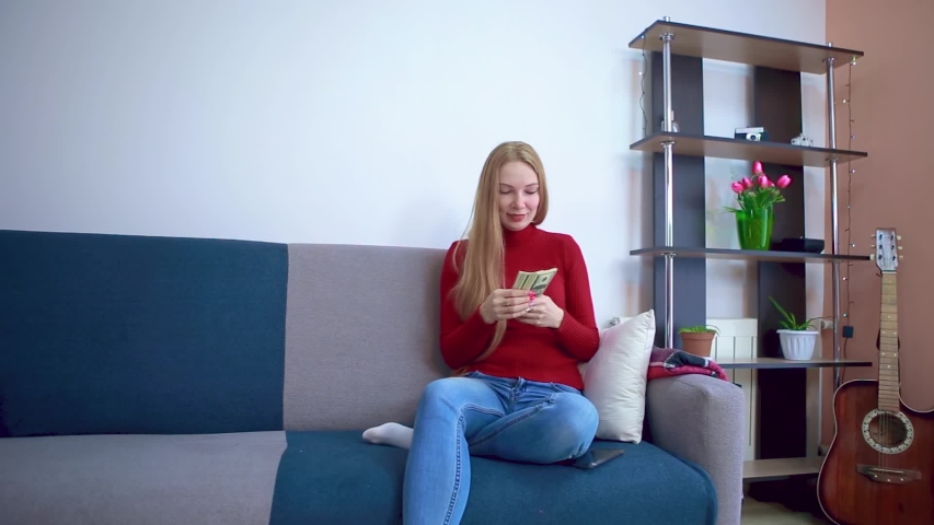 The girl in the red sweater sitting on the couch joyfully counts the money and thinks secretly about something good.
