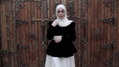 young lady model in traditional Arabian suit and white shayla decorated with pearls walks against wooden gate slow motion Video Stok