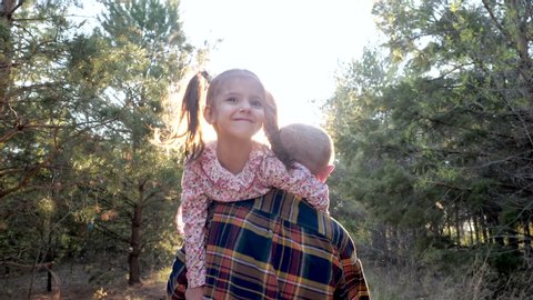 Back View at Old Grandfather Carrying Cute Smiling Child Girl on Shoulder, Walking in Summer Forest in Sunshine. Happy Lovely Family Plays Adventure Together, Fun Care for Little Grandchildren Outdoor
