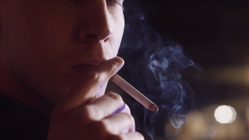 Close up of a young, handsome man smoking a cigarette in a dark alley. Lips blowing smoke with black background. Gangster during the night in a city. | Shutterstock HD Video #1053551894