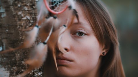 Portrait of beautiful young caucasian woman with green eyes posing in the forest near tree bark with dreamcatcher in formt of her eyes.