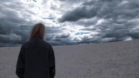 Blonde woman in depression is walking in a desert with clouds on background 