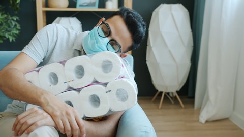 Portrait of crazy Middle Eastern guy in medical mask hugging pack of toilet paper rolls at home sleeping on couch after panic shopping because of covid-19 pandemic.