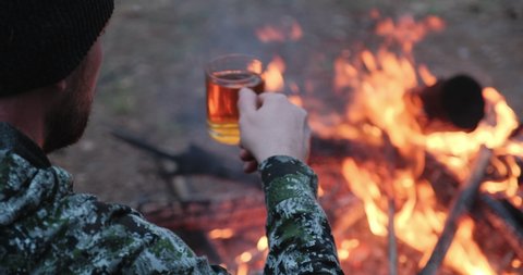 Man drinks hot tea near fire in the forest . Human looks at the fire and drinks tea on nature in the autumn. Rear view to alone traveler who sits, rests by the fire with a cup of coffee in evening.   Video Stok