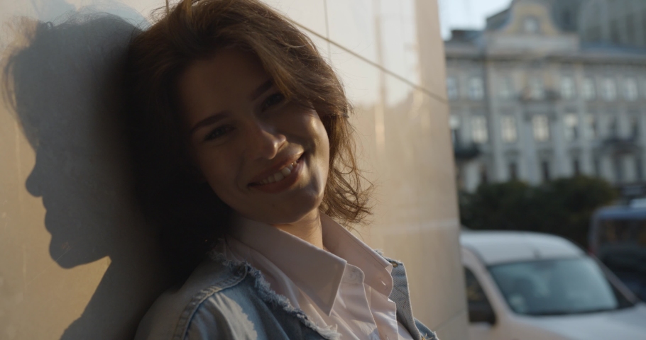 Attractive girl with short hair stands leaning against a wall in the street and smiles. girl on the wall shadow.
