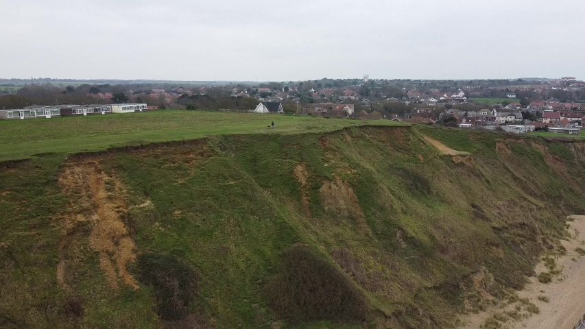 Footage of a distant person walking atop a cliff with their dog overlooking the beaches of norfolf with the town of Mundesley in the distance