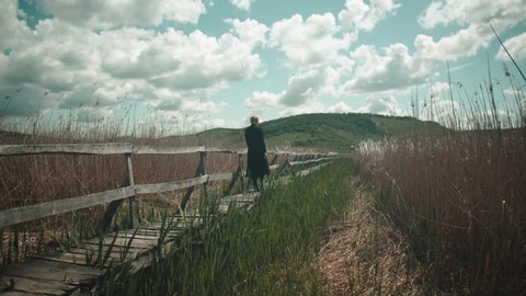 Slow motion shot of a beautiful blonde girl, dressed in black, walking on an old wooden deck over a reed bed