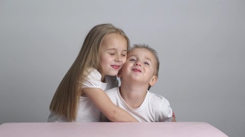 Caucasian little sister and brother in white T-shirts and blond hair sitting at the table. sister hugs brother and kisses