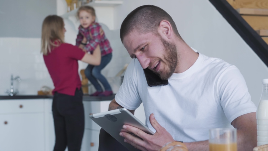 Busy young Caucasian man talking on the phone and using tablet as blurred wife and daughter having fun at the background. Portrait of brunette guy dealing with business on weekends. Lifestyle.
