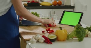 The woman is slicing cherry tomatoes and looking at the tablet. Tablet with green screen. Salad preparation. There are lots of vegetables around. 4K