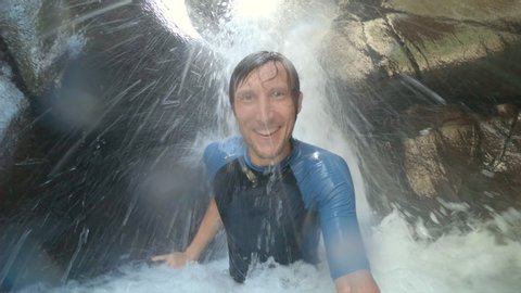 Slowmotion shot of a young man recording himself in a tropical river with a waterfall. Summertime concept