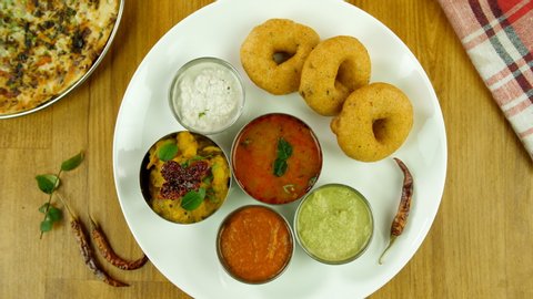 Top view shot of popular South Indian platter rotating on a wooden platform. Closeup shot of Sambhar Vada served in a ceramic white plate with colorful chutneys, potato masala, and Uttapam