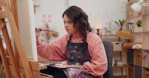 Pretty teenager girl wearing artist aprone soaking brush into paint. Adorable child with down syndrome holding art palette and painting while sitting in front of molbert in her room