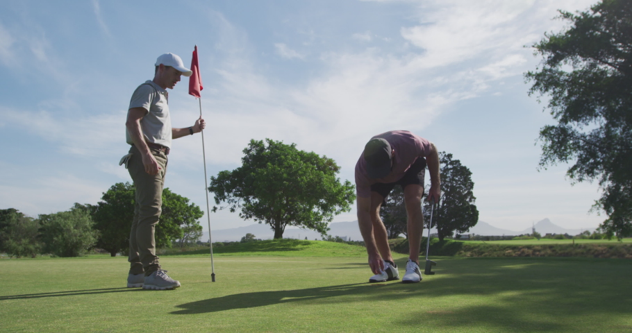 Two Caucasian male golfers on a golf course wearing caps and golf clothes on a sunny day, one player standing and holding the flagstick while his opponent takes his ball out of the final hole
