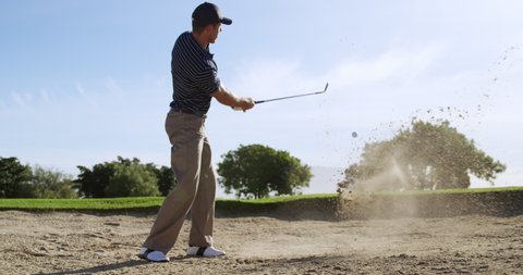 A Caucasian male golfer on a golf course on a sunny day wearing golf clothing, standing in a sand bunker, swinging a golf club and attempting to hit the ball out of the bunker towards the hole