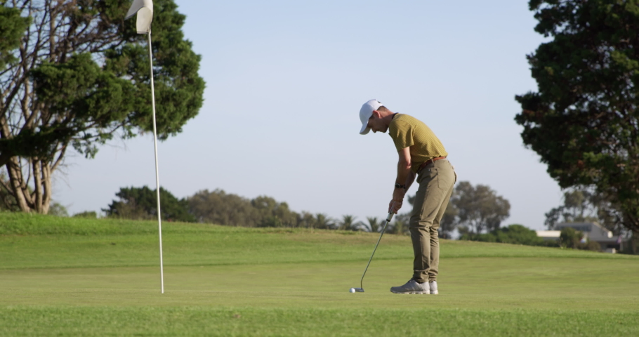 A Caucasian male golfer on a golf course on a sunny day wearing a cap and golf clothes, holding a golf club and putting the ball into the final hole, marked with a flagstick in the foreground Royalty-Free Stock Footage #1053605645