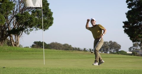 A Caucasian male golfer on a golf course on a sunny day wearing a cap and golf clothes, holding a golf club and putting the ball into the final hole, marked with a flagstick in the foreground