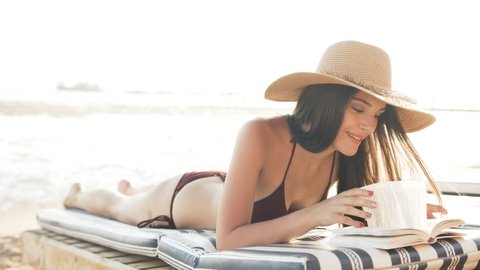 A woman is lounging on a sun bed by the sea and reading a book
