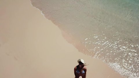 A drone shot of a girl with a straw hat walkin on the sand at the seashore of Pink Beach, Lombok, Indonesia. Waves gently washing the shore. Girl is suntanning. Happy moments.
