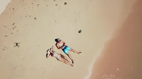 A couple playing with the drone while lying on the sand at the seashore of Pink Beach, Lombok, Indonesia. Waves gently washing the shore. They are suntanning. Happy and careless moments.
