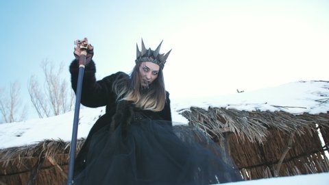 The witch in black robes with a crown looking into the camera in the winter against the background of wooden hut.