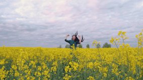 A girl against the background of the sunset dancing in a field of flowers. She has dark long hair and a long green dress. In the foreground are the bright flowers of rapeseed.