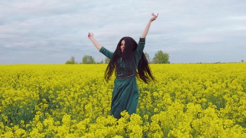 A brunette woman in a green dress is jumping and spinning in a blooming orange field. Girl's hands are raised to the sky. Long hair develops in the wind.