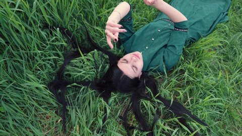 Beautiful girl in a green dress lies on the grass and rhythmically moves her hands. Looks up, brunette's hair is lying on the grass. Gnats fly. Mysticism, energy, meditation, occultism.
