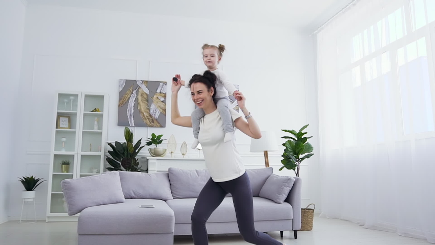 Beautiful slender positive happy mother doing squat exercises with her charming satisfied little girl on her shoulder in the living-room | Shutterstock HD Video #1053630626