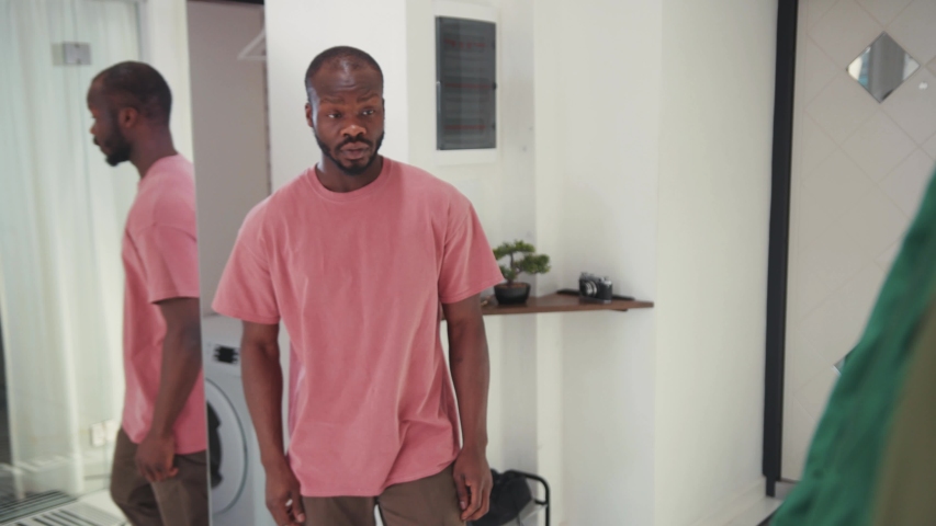 Young african american man puts on green jacket in front of mirror. Stylish guy getting dressed for work leaves the house going out. Casual people. Lifestyle. Apartment.