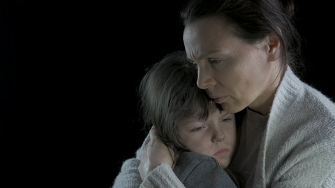 Concept of despair. Portrait of a homeless mother with a kid. Sad woman hugs a dirty little daughter on a black background.