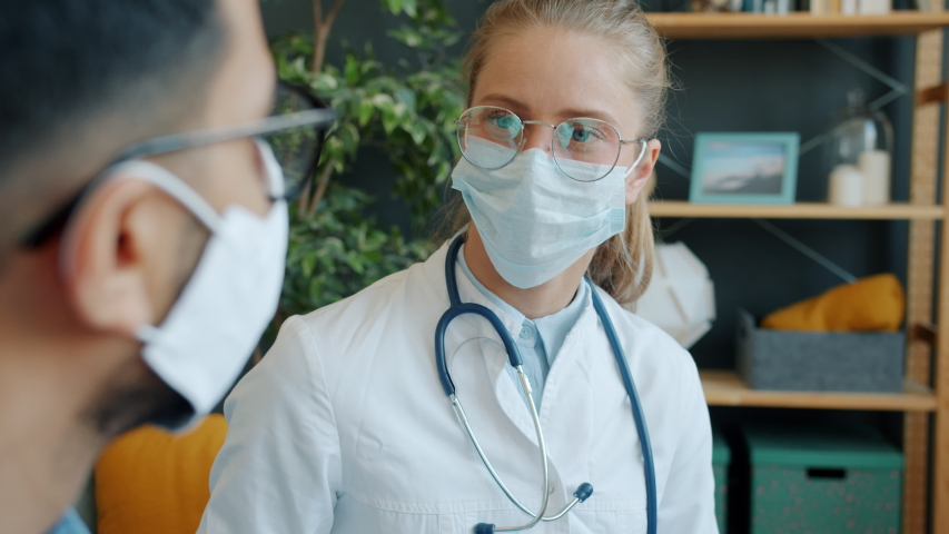 Young female doctor in face mask is talking to male patient at home while man is couching during covid-19 pandemic. Healthcare, medics and medicine concept. | Shutterstock HD Video #1053639761