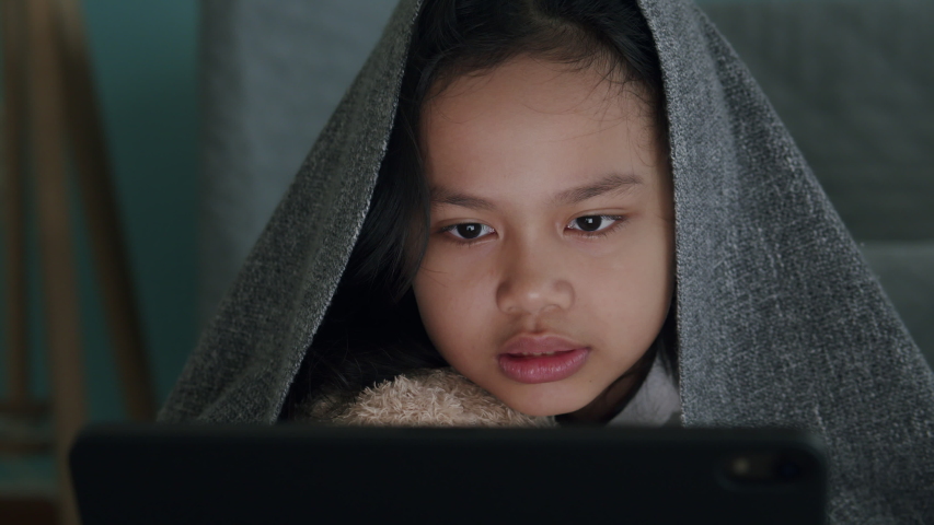 Asian little girl lying under blanket watch entertainment on a digital tablet in the bedroom at home night. Asia of child happy smile and hug dolly