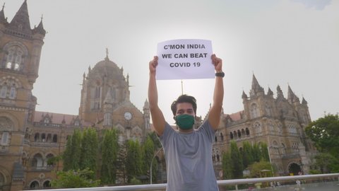 A young man wearing face mask standing and holding a placard with message 'C'mon India we can beat Covid 19' during city lockdown amid coronavirus corona against historic CSTM or VT terminus station
