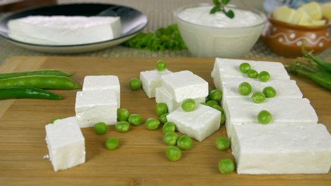 Closeup shot of organic green peas falling on the pieces of fresh white paneer. Shot of kitchen counter with slices of paneer on a chopping board surrounded by curd, butter, and peas - food concept