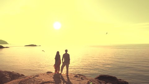 Happy Couple Silhouette standing on rock cliff toward to sea enjoying warm sunset with flying seagulls around