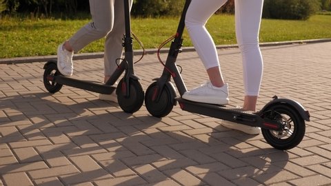 Close-up of teenagers riding electric scooters in the Park in the summer. Close-up of the legs of a guy and a girl in white sneakers on electric scooters.