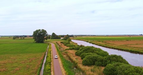AERIAL: White delivery van drives on a small road through green fields with a canal on the side in Friesland, the Netherlands 4K/UHD