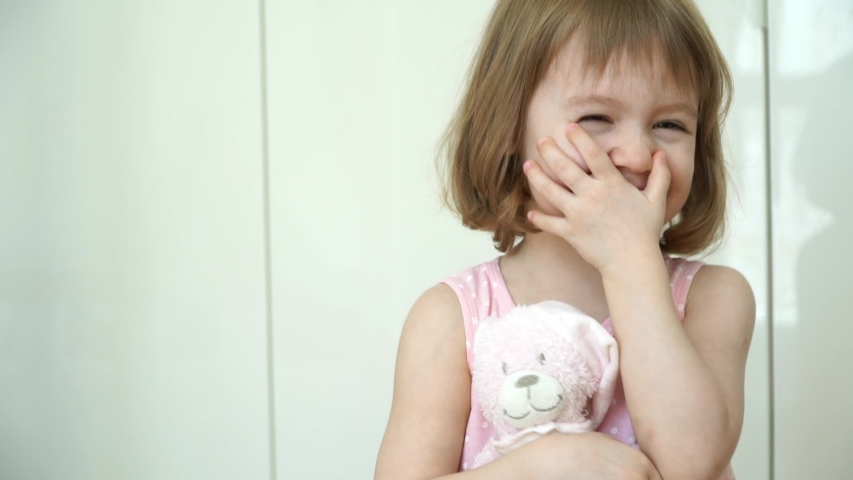 Portrait of little cute funny 3 year old girl looks at camera, smiles and laughs. Happy child plays with hands and holds in hands pink teddy bear. Close up Royalty-Free Stock Footage #1053653609