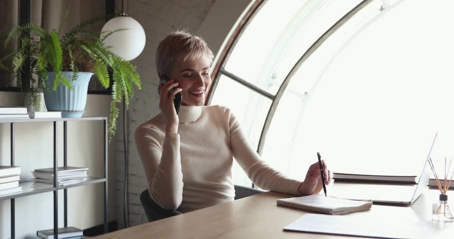 Smiling young business woman talking on mobile phone sitting alone at desk in modern office. Female professional manager entrepreneur making business call having mobile conversation at workplace. Royalty-Free Stock Footage #1053657626