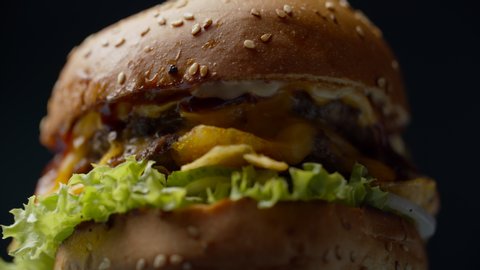 Yummy burger with double beef cutlet, melted cheese, lettuce and vegetables rotating on dark background in slow motion.