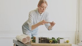 A happy successful young blonde woman architect wearing glasses is taking a top view photo of model-house while sitting in the modern office