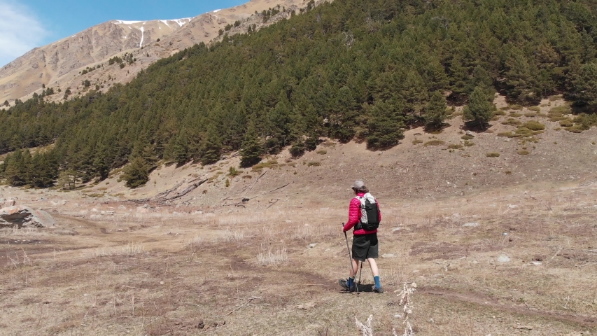 Aerial view of a stylish young man in a sunglasses cap and shorts with a jacket walks trekking with sticks along the gorge