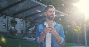 Young Handsome Man Standing at the Bus stop with Mobile Phone, Using social Networks. Chatting in the online Conversation. Having fun and smiling Sincerely. Casual Lifestyle. Phones. Emotions.
