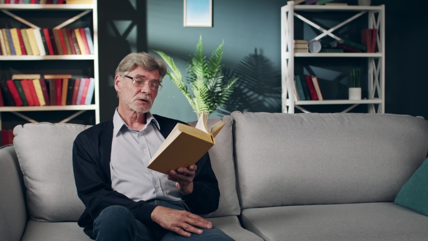 An elderly grey man in glasess is sitting on a couch and thinking over the content of the book he is reading. An old man is reading a book out loud in a cozy living room.