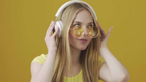 Young Blonde Girl in Yellow Glasses, Yellow T-shirt and White Headphones Listening to Music and Dancing on Yellow Background. Portrait of Beautiful Girl in Round Glasses on Yellow Background in Studio